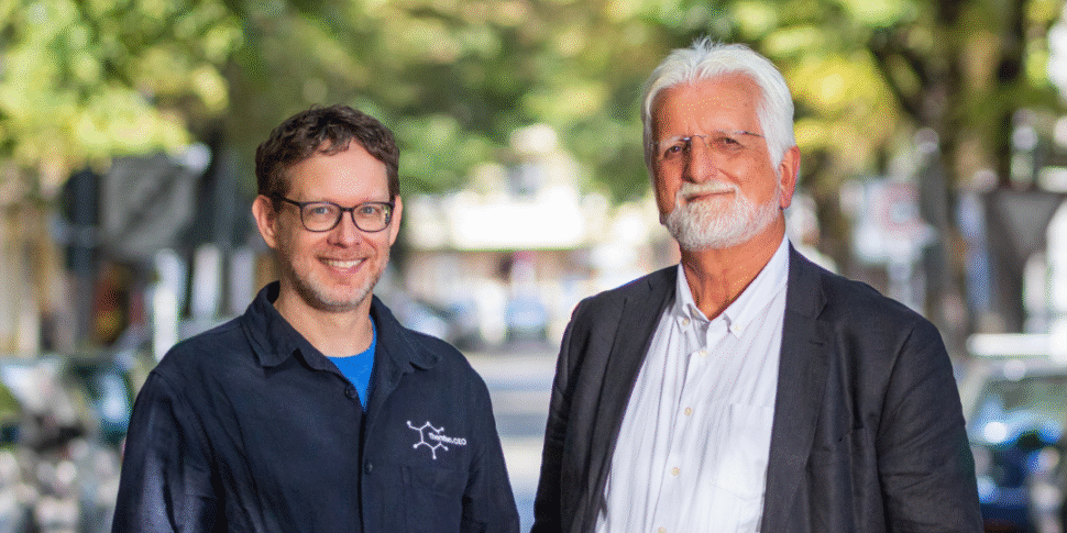 Zwei Männer, die lächelnd in die Kamera schauen. Im Hintergrund sind Bäume zu erkennen.