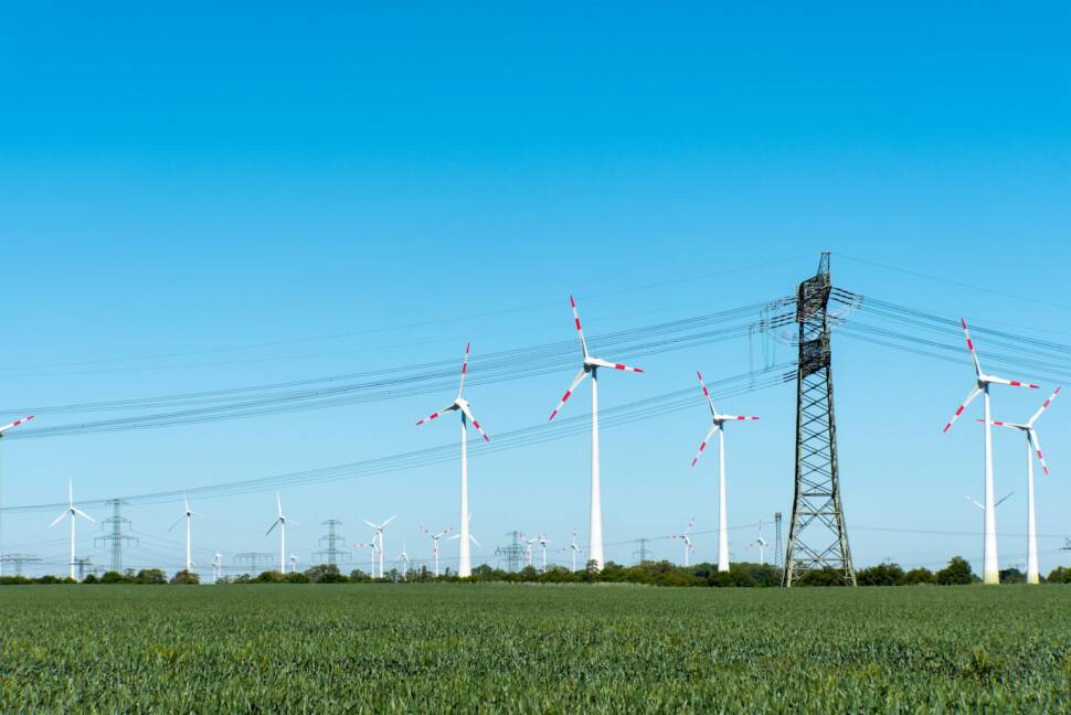 Landschaft mit Feldern und Strommast im Vordergrund und Windrädern im Hintergrund