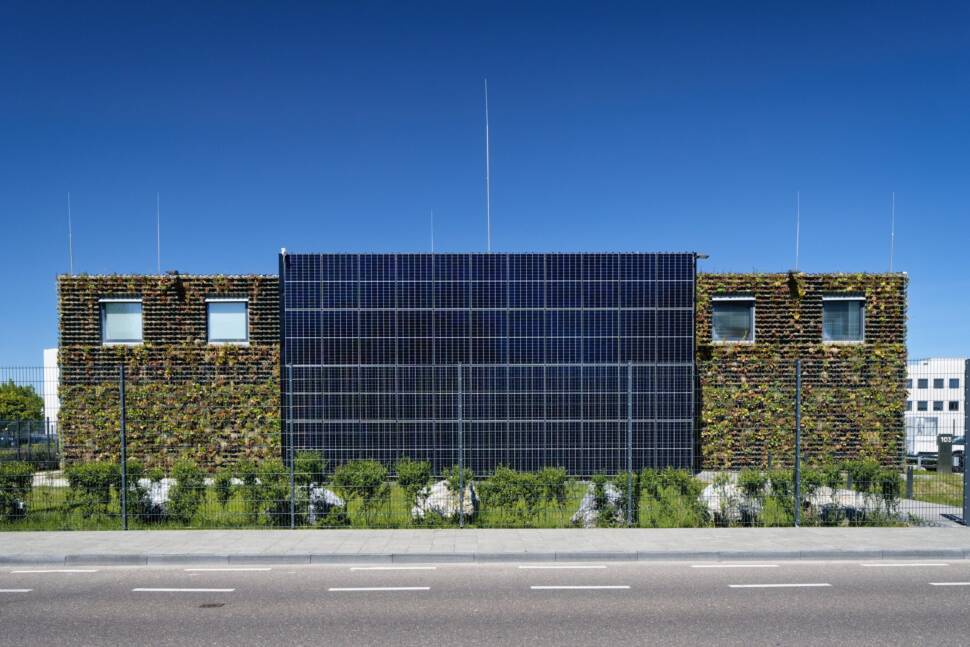 Flachdachgebäude mit begrünter Wand und Solaranlage an der Wand hinter einem hohen Zaun.