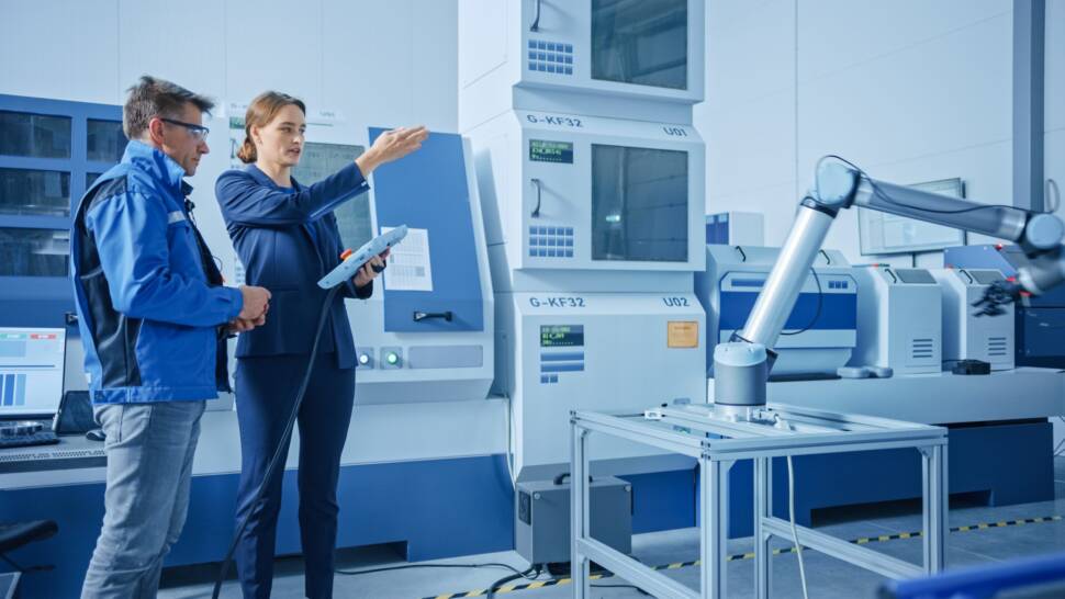 Moderne Fabrik: Projektmanagerin und Ingenieurin stehen, sprechen und verwenden Tablet-Computer zur Programmierung von Roboterarm-Montagelinie.
