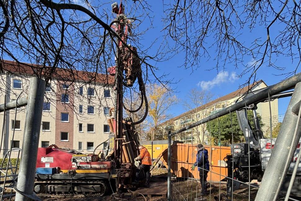Bohrgerät mit Arbeitern, Bauzaun und einem Container, Wohngebäuden im Hintergrund