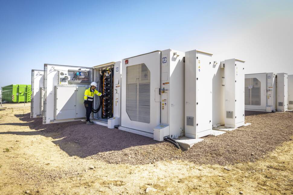Große weiße Container auf einem Feld. Ein Arbeiter in Schutzweste hat die Tür des vorderen Containers geöffnet. Es sind große Kabel zu erkennen.
