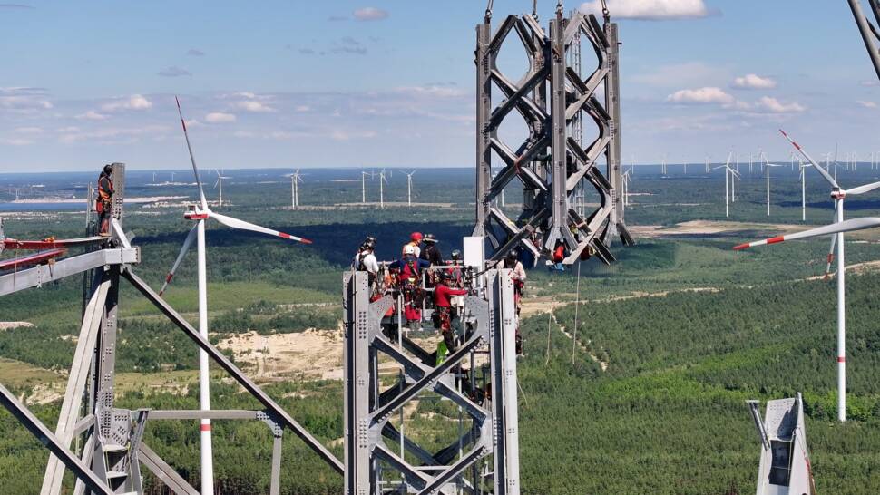 Montage eines Mastelements in großer Höhe, mehrere Arbeiter mit höhensicherungsausrüstung warten, im Hintergrund (unterhalb) sind andere Windräder zu sehen
