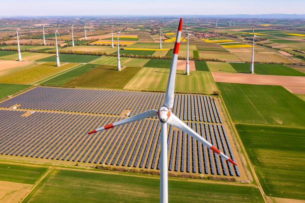 Luftaufnahme auf eine Landschaft mit vielen Feldern, Wiesen. Vorne ein großer Solarpark auf Feldern, im Vordergrund eine Windkraftanlage, im Hintergrund viele weitere.